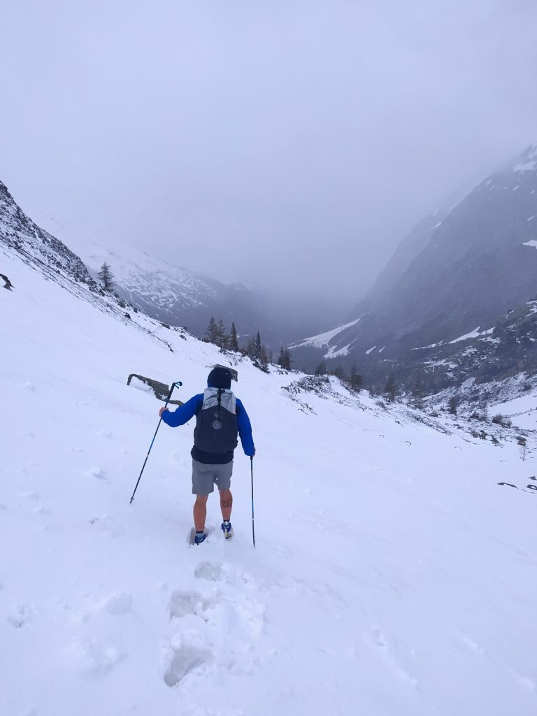 Hiker in shorts on snow in the mountains on a gray day, using long hiking poles on TMB hiking trail in Val Ferret